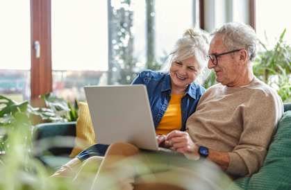 An older couple is sitting on a couch, looking at a laptop together.