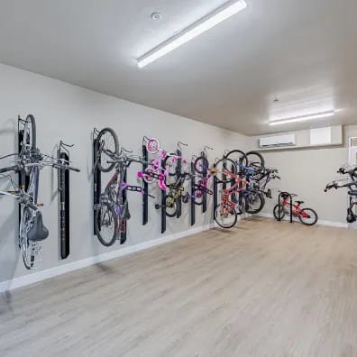 A room with a white wall and a wooden floor is filled with bicycles of various colors and sizes.