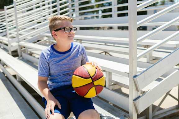 A boy with glasses is sitting on a bleachers holding a basketball.