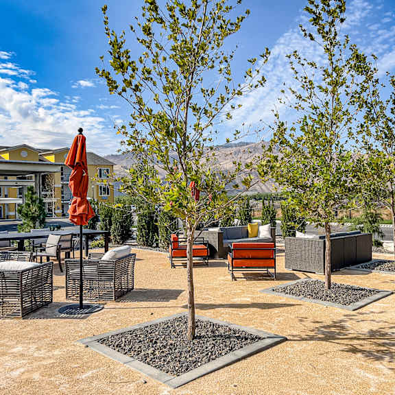 A courtyard with plush chairs, tables, umbrellas, and trees, with the apartment buildings and mountains in the background at LIVEasy Apartments. 