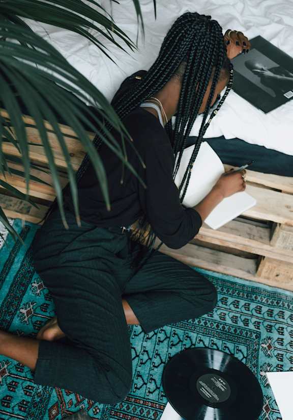 A person with braided hair is sitting on a patterned rug, writing in a notebook with a pen, and there is a vinyl record and a book beside them.