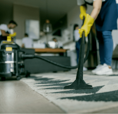 A person in yellow gloves is vacuuming a rug in a room with other people in the background.