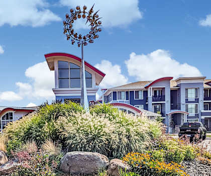 The roundabout with a metal sun sculpture in the middle surrounded by plants with the unique white, blue, and red leasing office in the background at Solara Apartment Homes. 