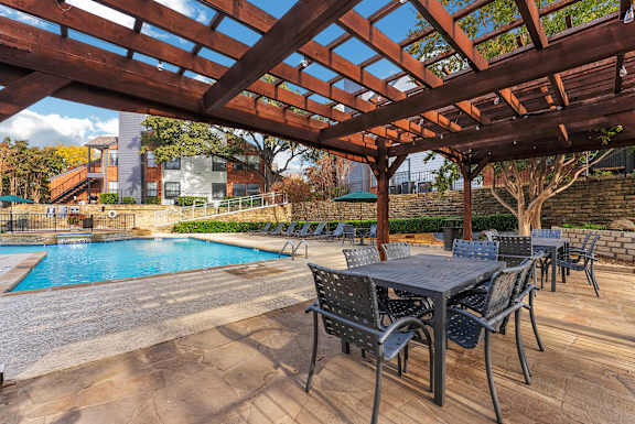 A wooden pergola over a table with chairs and a pool in the background.