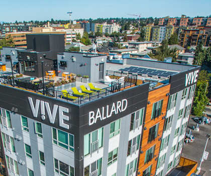 An arial view of the Vive white, gray, orange, and black apartment building with the rooftop lounge with string lighting, couches, chairs, coffee tables, and potted plants. 