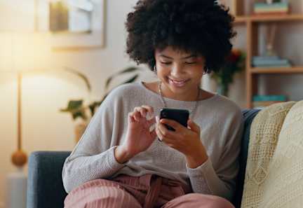 A woman with curly hair is sitting on a couch and looking at her phone.