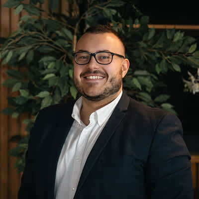 A man with glasses and a beard is smiling in front of a green plant.