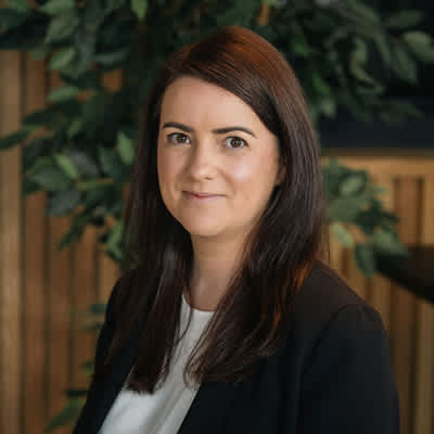 A woman with dark hair and a white shirt is standing in front of a wooden wall with green leaves.