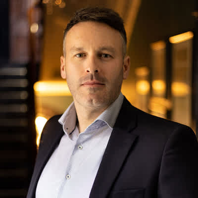 A man in a blue shirt and black jacket stands in front of a blurred background.
