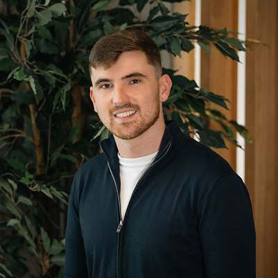 A man with a beard and short hair wearing a black zip-up hoodie stands in front of a green plant.