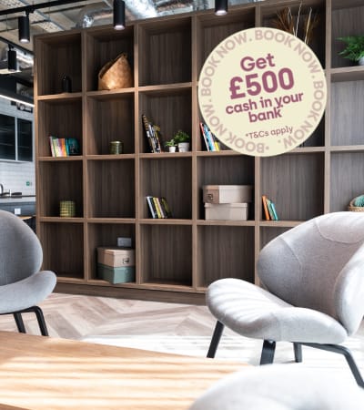 A room with a grey chair, a wooden table, and a bookshelf with a heart-shaped sticker on it.