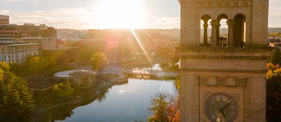 A clock tower stands tall in the foreground of a serene landscape with a river and buildings in the background.