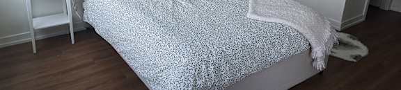 A bed with a grey and white patterned duvet cover sits in a room with a white nightstand and a lamp.