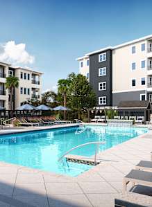 A swimming pool surrounded by sun loungers and umbrellas in front of apartment buildings.