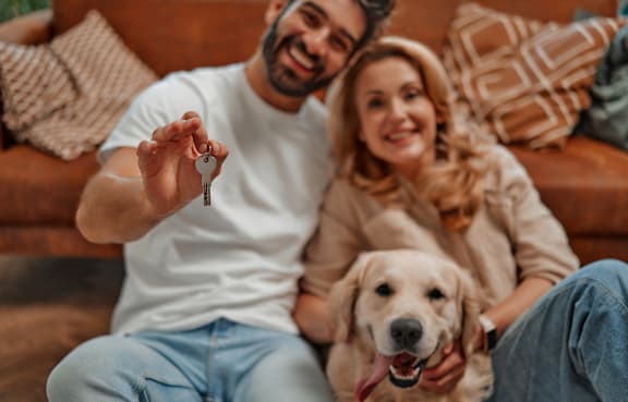 A man and woman are sitting on a couch with a dog, and the man is holding a key.