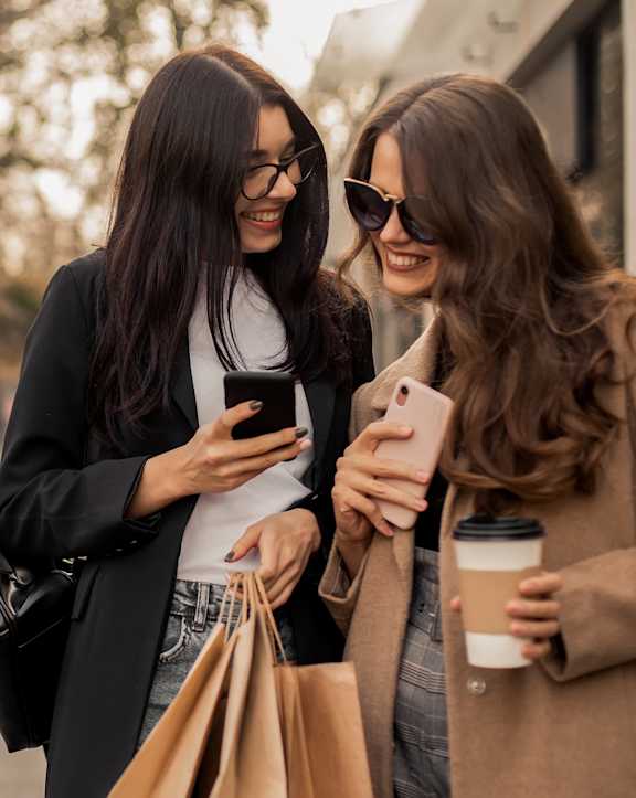 Two women are standing outside, one holding a phone and the other holding a coffee cup.