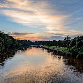 A serene river flows through a lush green landscape under a sky painted with hues of sunrise or sunset.