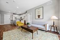 A living room with hardwood-style flooring, natural light, high ceilings, and open access to the kitchen with a breakfast bar.