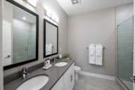 A bathroom with double vanity sinks, two large mirrors, a toilet, and a swinging glass door to a stand up shower with tile walls.