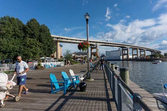 A man pushing a stroller walks on a pier with blue chairs and a bridge at The Lofts at Elk Terminal Apartments, Buffalo, NY