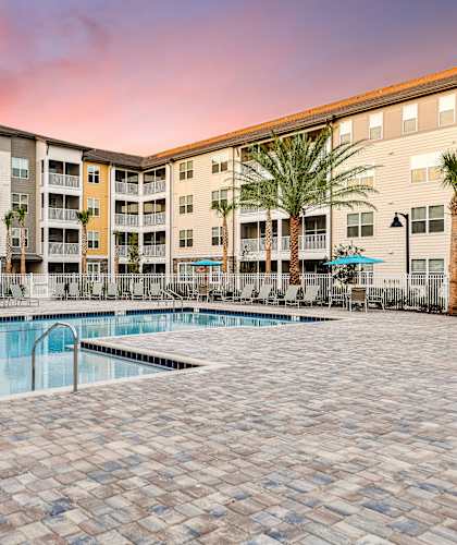 A large swimming pool surrounded by palm trees and lounge chairs in front of a multi-story apartment building.