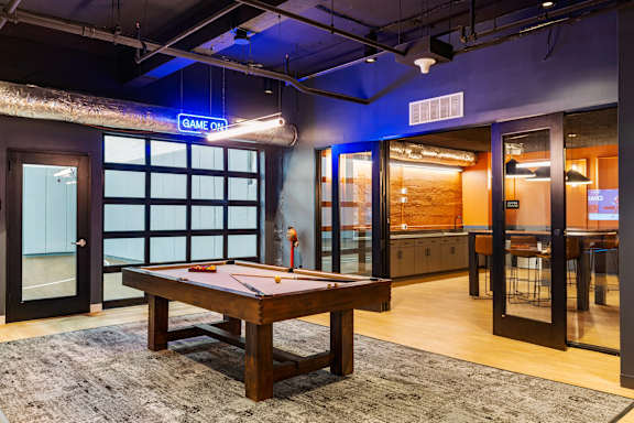 A pool table is in the middle of a room with a blue neon sign that says "GAME ON".