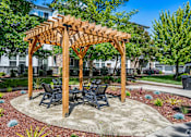 Thumbnail 5 of 20 - A wooden pergola with a table and chairs is surrounded by a gravel area with small plants.