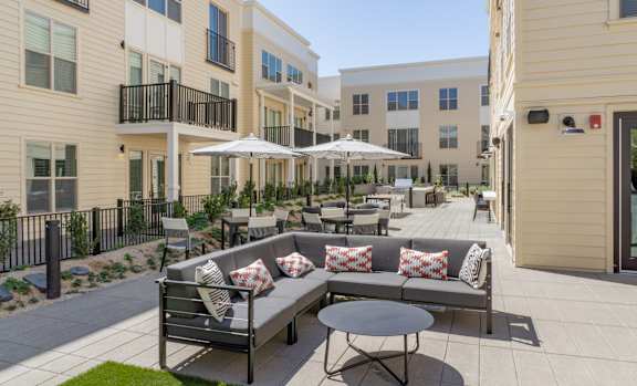 A patio with a couch, chairs, and umbrellas is surrounded by apartment buildings.