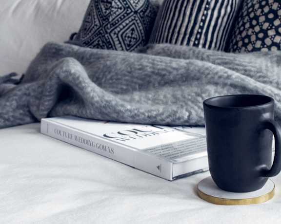 A black mug sits on a cozy bed with a book titled "Color Wedding Gowns".