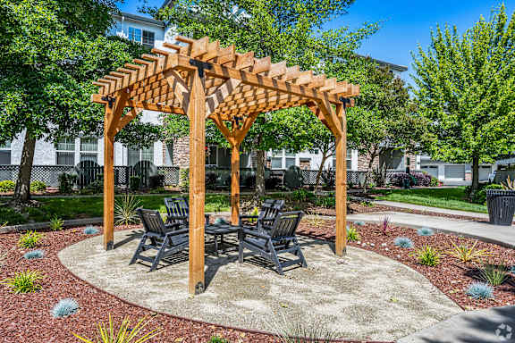 A wooden pergola with a table and chairs is surrounded by a gravel area with small plants.