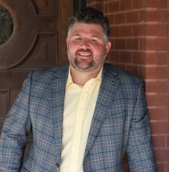A man in a black suit and white shirt stands in front of a red brick wall.