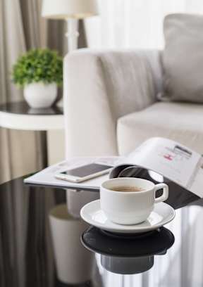 A cup of coffee sits on a saucer with a newspaper and a smartphone on a table.