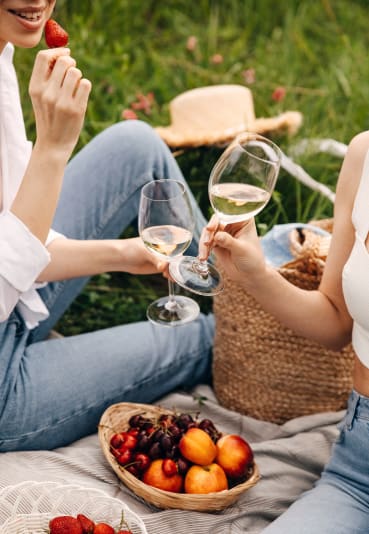 Two people are enjoying a picnic in a grassy area, with a basket of fruit, two glasses of white wine, and a hat visible. The person on the left is eating a strawberry.