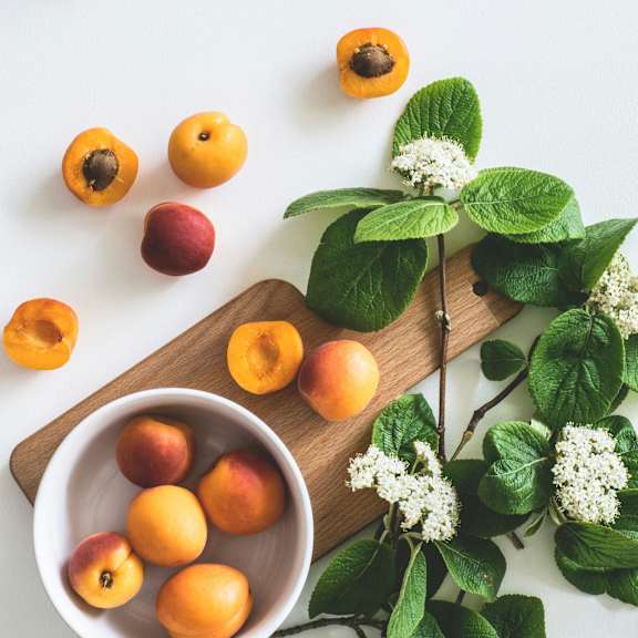 A bowl of fresh apricots on a wooden board with some leaves and flowers.