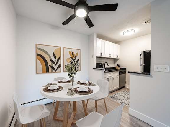 A dining room with a white table and chairs, a ceiling fan, and a kitchen area in the background.