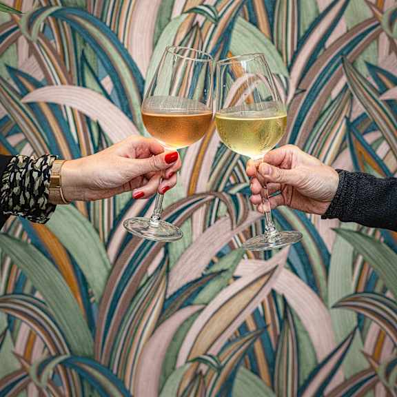 Two hands toasting with wine glasses in front of a colorful wallpaper.