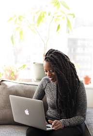 A woman with dreadlocks is sitting on a couch using a laptop.