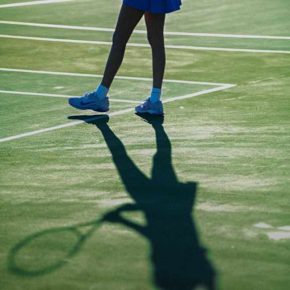 A tennis player is captured in mid-swing, casting a long shadow on the court.