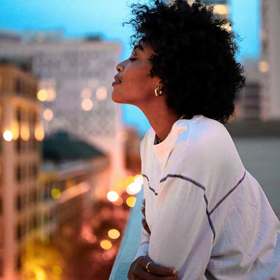 A woman with curly hair is standing on a balcony looking at the city lights.