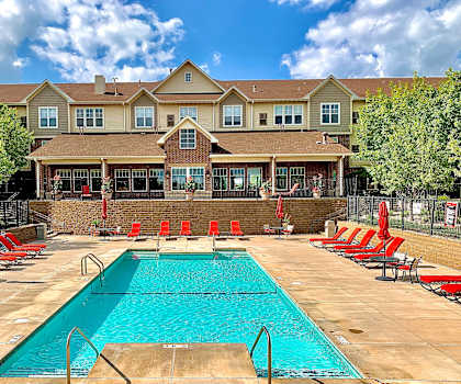 The large, turquoise outdoor pool with red pool chairs and red umbrellas with the apartments in the background at Stoneleigh at the Reserve Apartment Homes. 