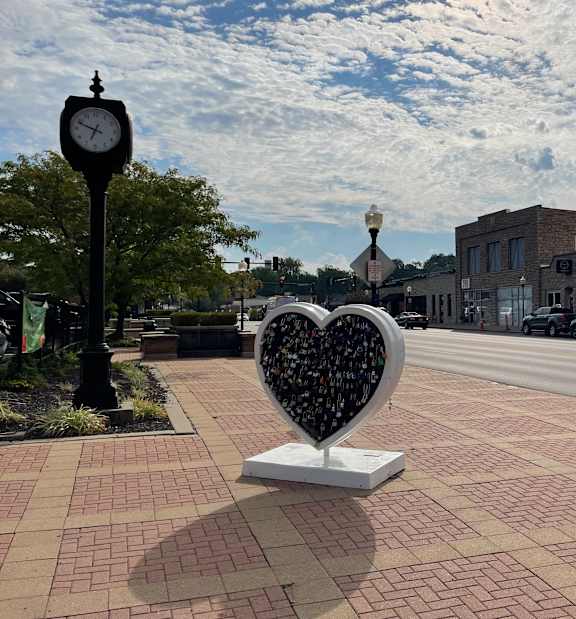 A heart-shaped sculpture made of small objects is in the foreground of a city street.