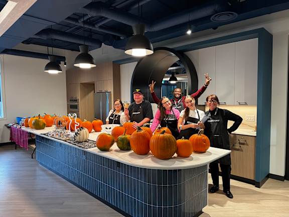 A group of people in a kitchen with pumpkins on the counter.