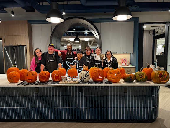 A group of people in a kitchen with carved pumpkins in front of them.