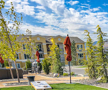 A courtyard with a game of cornhole and grass, bushes, trees, the buildings, and hills in the background at LIVEasy Apartments. 
