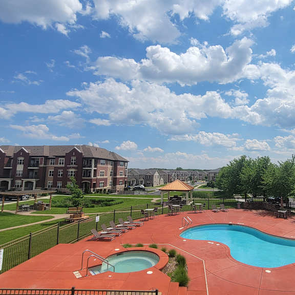 The large, turquoise outdoor pool with pool chairs, tables, and chairs on a red clay concrete, with the green space and apartments in the background at Van Buren Place Apartment Homes. 