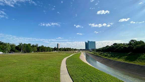 A river flows through a green field with a road and buildings in the background.
