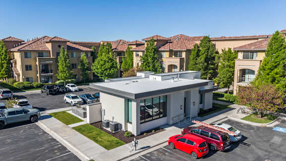 A red car is parked in front of a white building with a parking lot in front of apartment buildings.