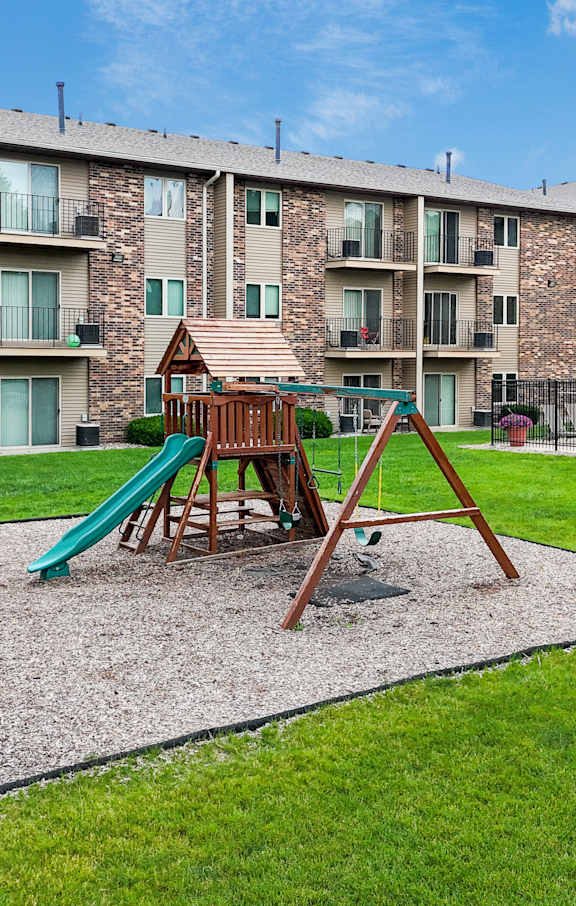 A playground with a slide and swing set in a grassy area in front of apartment buildings.