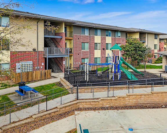 A playground area with a slide and a play structure is surrounded by a fence and apartment buildings.