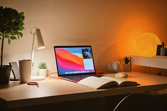 A laptop is open on a desk with a book and a lamp beside it.
